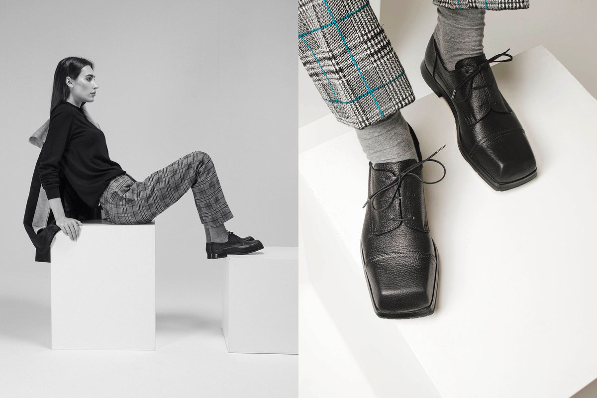 Editorial still life and on-foot detail of black leather lace-up shoes featuring a square toe, paired with checked tailored trousers and grey socks. Minimalist black-and-white setting with a model seated on white geometric blocks.