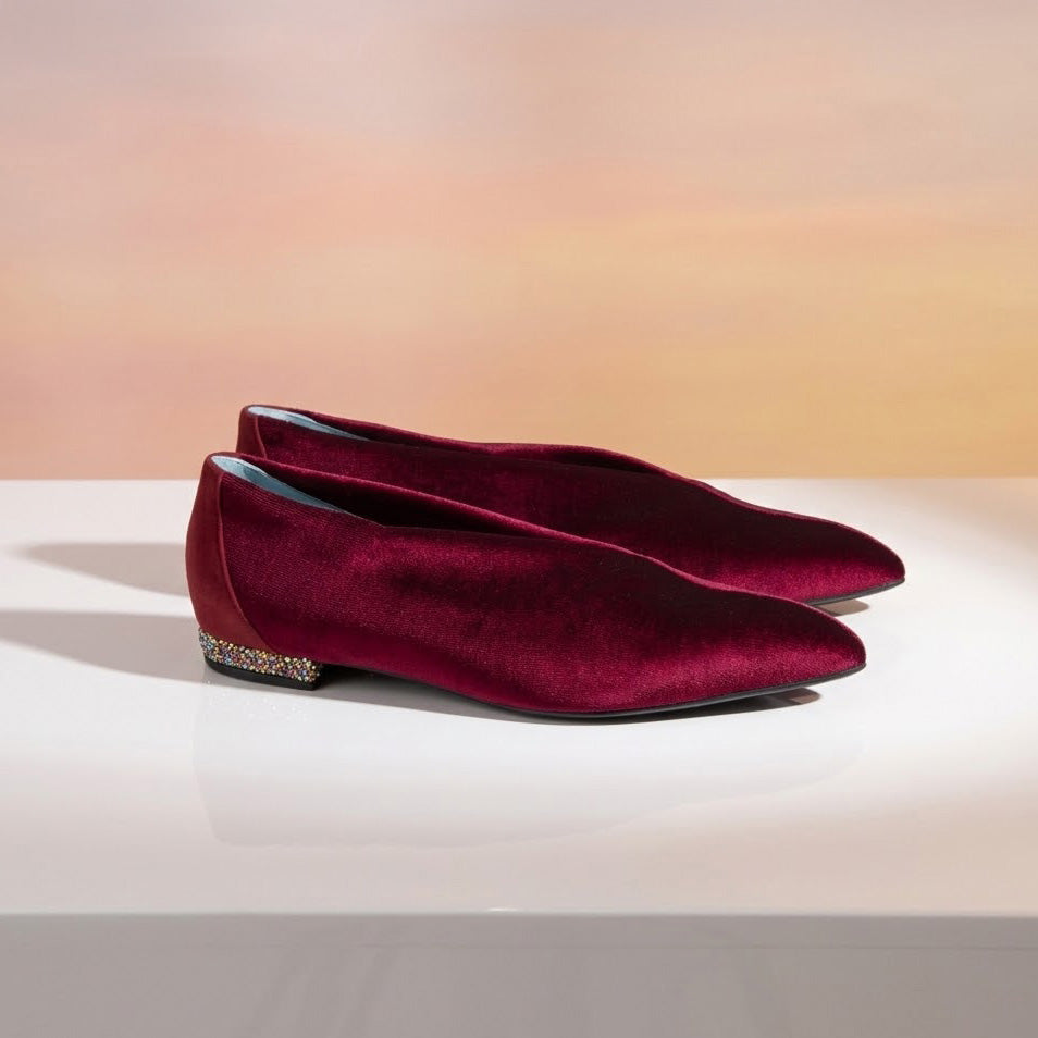A pair of deep burgundy velvet pointed-toe flats with a jeweled embellished heel, displayed on a white surface against a warm peach gradient background.